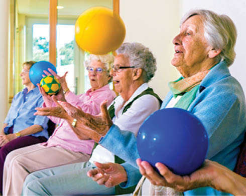 Group of happy senior ladies doing coordination exercises in a seniors gym sitting in chairs throwing and catching brightly colored balls.